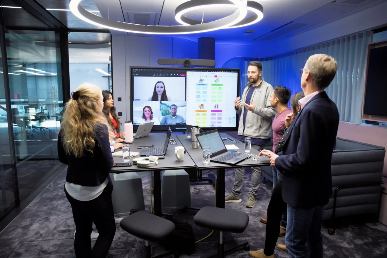 SAP-Mitarbeitende stehen an einem großen Tisch in einem Meetingraum. Auf einem mobilen Bildschirm sind Mitarbeitende zugeschaltet. Eine Person in der Gruppe spricht gerade.
Foto: SAP SE